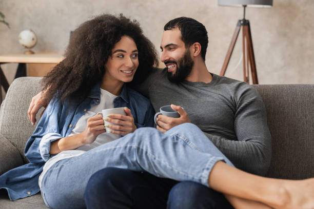 A young biracial couple is cuddled together on the couch, drinking coffee.