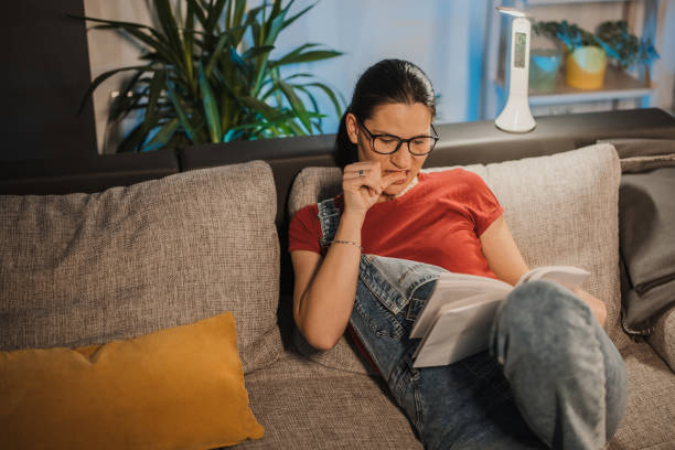 A young woman is sitting on her couch in the living room reading a book, as she bites the skin on her thumb.