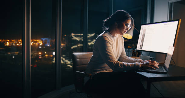 A young business woman is sitting in her office at night. She is typing on her laptop.