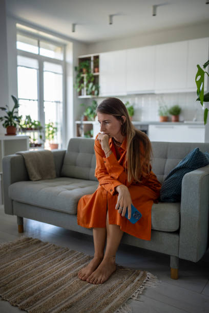 A young woman is sitting on her couch in her living room, with her hand resting on her knees.