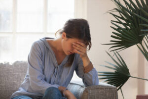 A young woman is sitting on the couch in her living room, with her head down and her hand on her face.