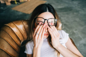 A young woman is laying down on a leather couch, with her glasses moved up as she rubs her eyes.