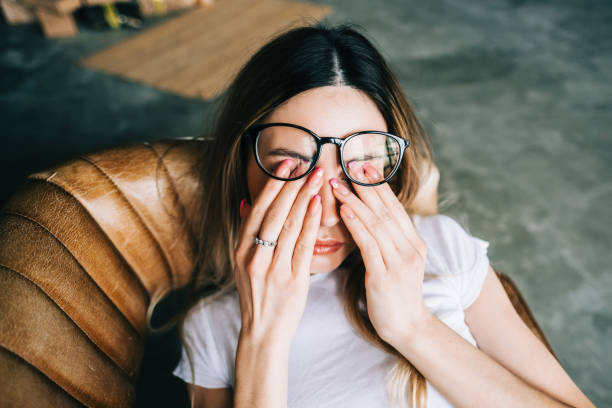 A young woman is laying down on a leather couch, with her glasses moved up as she rubs her eyes.