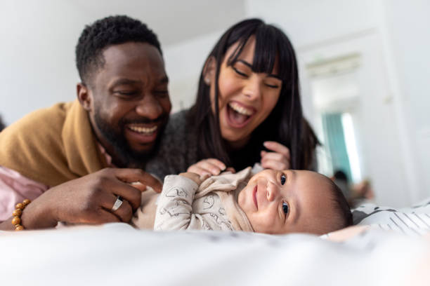 A biracial couple is laying on their bed, playing with their baby as they smile.