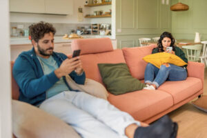 A young American couple is sitting on opposite ends of the couch. They are both preoccupied with their phones.