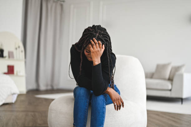 A young African American woman is sitting on a couch chair, with her hand on her head leaning forward.