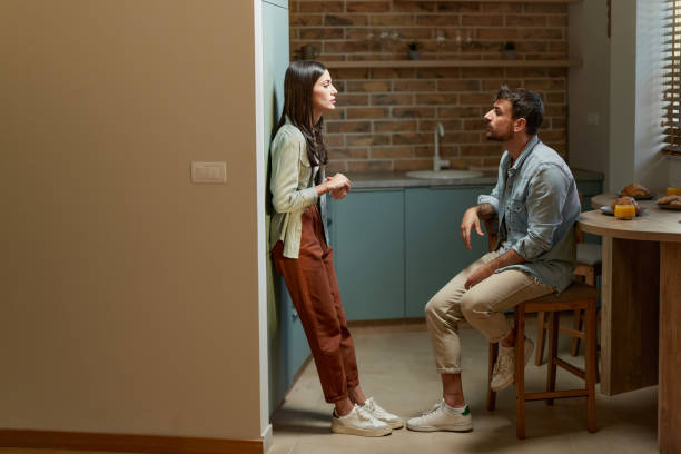 A young couple is in their kitchen having a conversation. The husband is sitting across from his wife on a chair, as she leans on the wall.