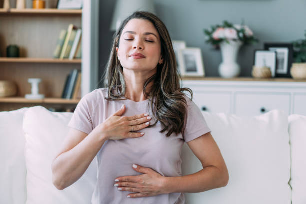 A woman is sitting on her couch with her eyes closed and her hands on her chest as she takes a breath in.