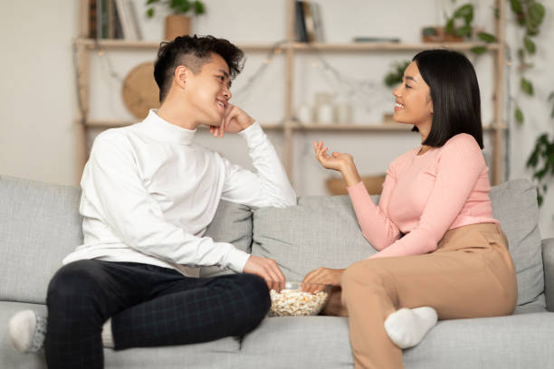 An Asian American couple is sitting together on the couch, talking and eating popcorn.