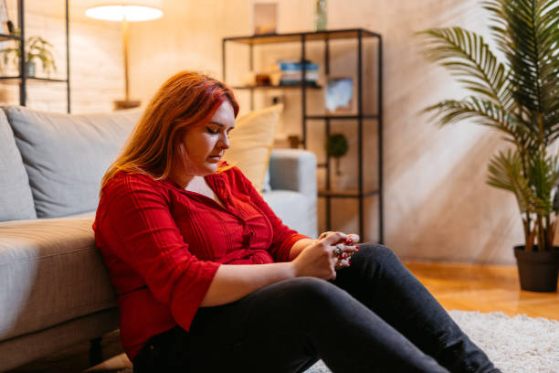 A young woman is sitting on the floor of her living room, leaning on her couch with her eyes closed.