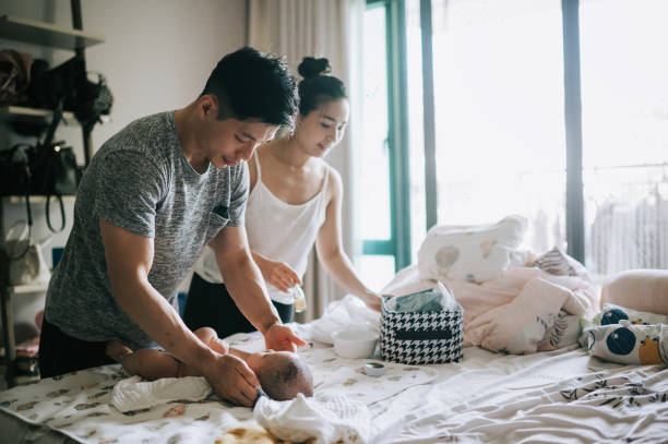 An Asian American couple is in their bedroom, changing their baby on the bed. The father is changing the babys diaper as the mother helps him.