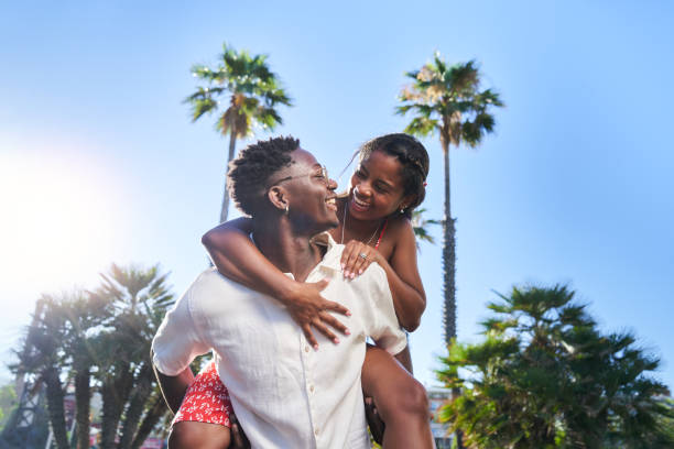 An African American couple is outdoors together, as the man carries the woman on his back.