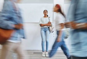 A young woman stands alone against a wall, holding a backpack and looking up pensively while people blur past her, emphasizing a sense of isolation or anxiety in a busy environment.