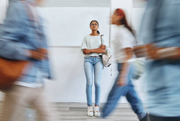 A young woman stands alone against a wall, holding a backpack and looking up pensively while people blur past her, emphasizing a sense of isolation or anxiety in a busy environment.