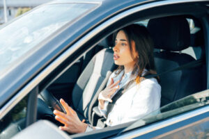 A young woman sitting in the driver’s seat of a car, looking anxious and holding her chest with one hand while the other rests on the steering wheel.