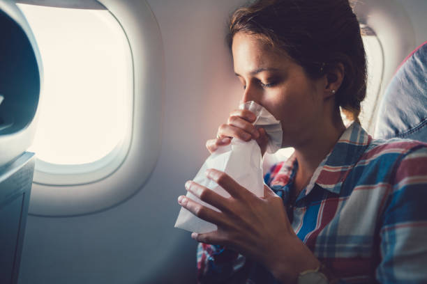A young woman is sitting on a flight, leaning on the window as she closes her eyes and breathes into a paper bag.