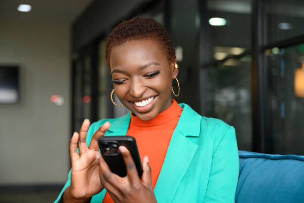 A young African American woman is sitting in her office smiling at her phone as she scrolls.
