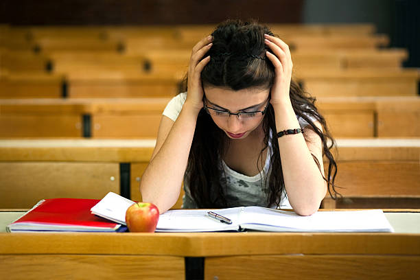 A stressed college student sits alone in an empty lecture hall, holding her head in frustration while studying with open books and an apple in front of her.