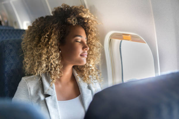 An African American woman is sitting in the window seat of a plane with her eyes closed.