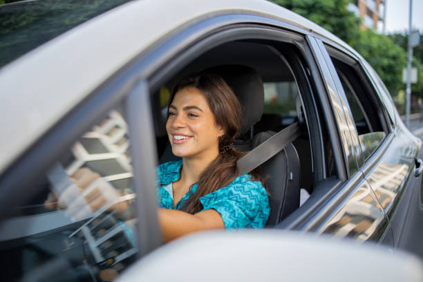 A smiling young woman driving a car with the window down, appearing relaxed and happy behind the wheel.