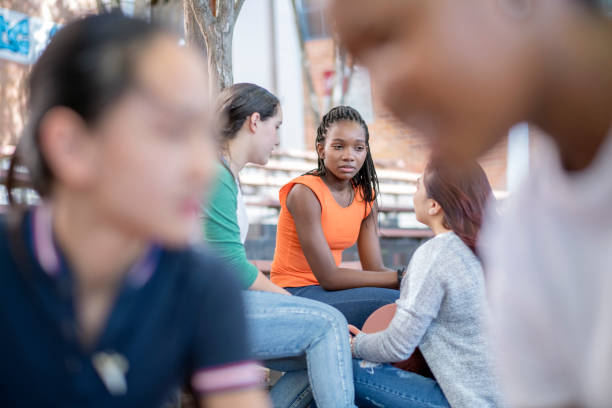 A teenage girl sits quietly among a group of peers, looking uncomfortable and isolated while others talk around her, capturing the experience of social pressure or exclusion.
