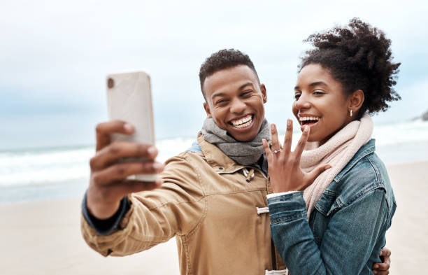 A joyful couple takes a selfie on the beach, with the woman proudly showing off an engagement ring, both smiling and bundled up in warm clothing, capturing a celebratory moment.