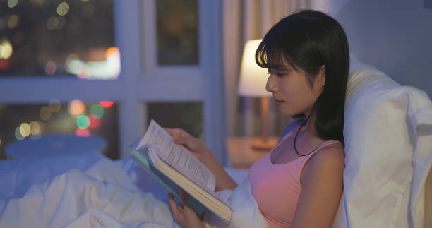 A woman sits in bed at night reading a book under soft lamp light, creating a calm bedtime routine in a cozy, dimly lit room with city lights outside the window.