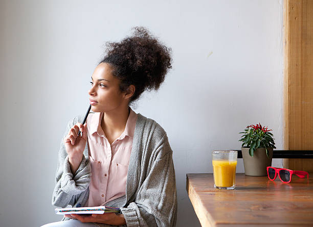A thoughtful woman sits at a wooden table with a notebook and pen in hand, gazing out of frame while appearing deep in reflection, with a glass of orange juice and a small plant beside her.