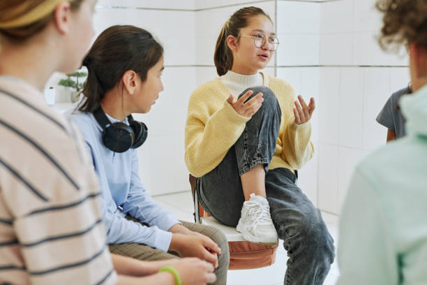 A group of teens sit in a circle having a serious discussion, with one girl in a yellow sweater speaking while the others listen attentively, suggesting a supportive peer conversation or group therapy setting.