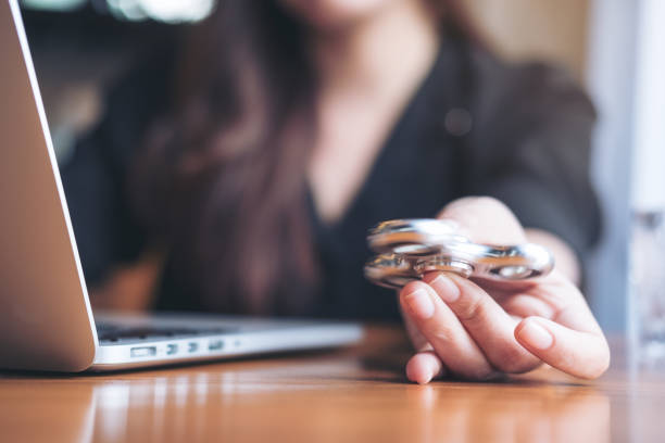 A woman sits at a desk in front of a laptop, holding a metallic fidget spinner in her hand, suggesting a moment of stress relief or focus aid during work or study.