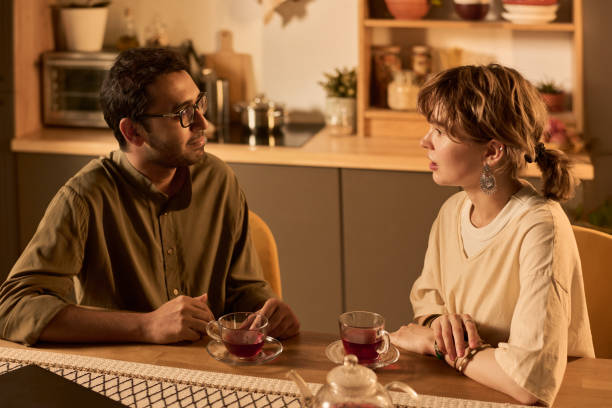 A couple sits at a cozy kitchen table, engaged in a thoughtful and serious conversation over cups of tea, suggesting an intimate and meaningful dialogue.