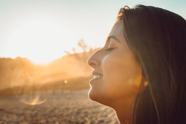 A woman with closed eyes smiles peacefully while facing the sun on a beach, capturing a moment of calm, mindfulness, and emotional well-being.