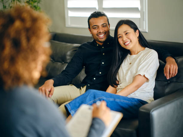 A smiling couple sits closely together on a couch during a counseling session, looking at a therapist who is taking notes, suggesting a positive and supportive atmosphere in premarital therapy.
