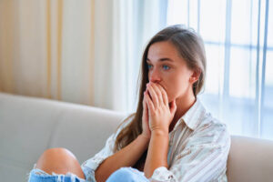 A young woman sits on a couch with her hands covering her mouth, looking anxious and deep in thought, conveying feelings of worry or emotional distress.