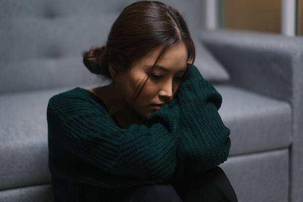 A young woman sits on the floor with her arms wrapped around her knees, looking down with a sad and withdrawn expression, conveying symptoms of adult depression or emotional overwhelm.