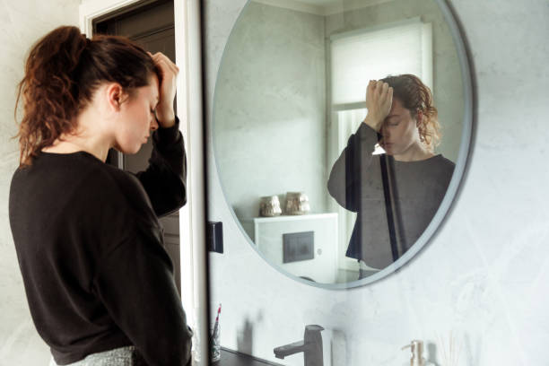 A woman stands in front of a bathroom mirror with her hand on her forehead, appearing distressed and deep in thought, reflecting feelings of self-criticism or body image struggles.