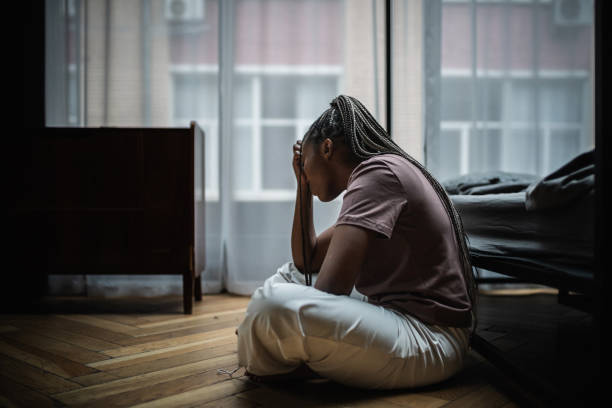 A woman sits on the floor in a dimly lit bedroom, holding her head in her hand with a posture of deep distress, symbolizing the emotional weight of adult depression or mental exhaustion.