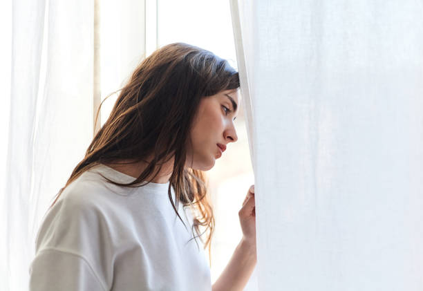 A woman stands quietly by a window, gently holding a curtain and looking outside with a pensive expression, conveying feelings of sadness, isolation, or self-reflection.