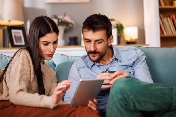Beautiful young couple in love sitting on living room couch, paying bills and taxes online using tablet computer and drinking coffee while relaxing and enjoying leisure time at home