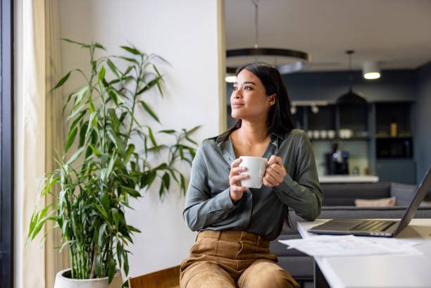 A woman sits by a desk holding a mug, gazing peacefully out the window with a soft smile, suggesting a moment of reflection, calm, or emotional recovery from a difficult time.