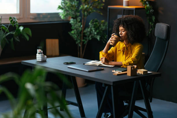 A woman sits calmly at a desk in a cozy, plant-filled office, eyes closed and holding a cup with both hands, suggesting a moment of mindfulness, self-compassion, or quiet reflection.