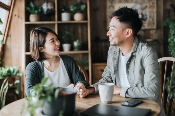 Happy young Asian couple having a coffee date in cafe, holding hands on coffee table. Drinking coffee and chatting. Enjoying a relaxing moment together