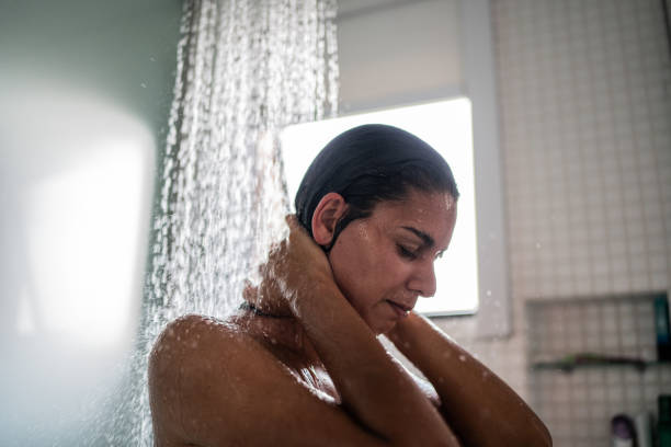 A woman stands under a shower with her eyes closed and hands on her neck, appearing calm and introspective, capturing a private moment of self-care or emotional release.