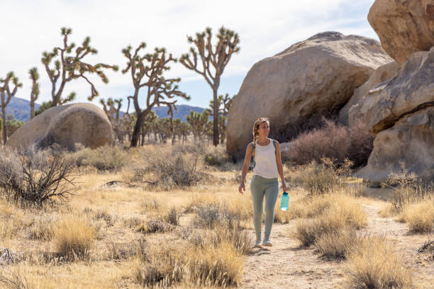 A woman walks alone through a desert trail surrounded by Joshua trees and large rock formations, holding a water bottle and appearing calm and reflective, suggesting self-care and solitude in nature.