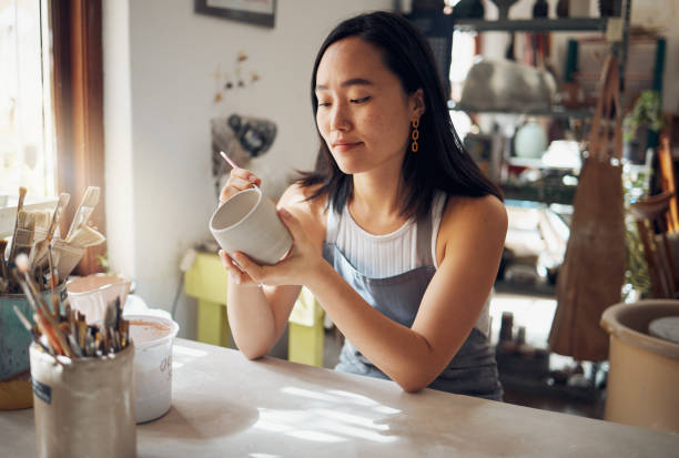 A woman sits at a table in a sunlit studio, peacefully painting a ceramic mug with focused attention, symbolizing creativity, self-expression, and self-care through art.