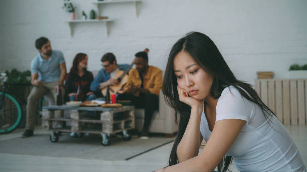 A young woman sits apart from a group of people socializing in the background, looking down with a sad and withdrawn expression, reflecting feelings of social anxiety or isolation.