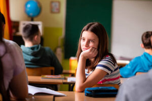 A young girl sitting in a classroom, appearing distracted and thoughtful while lightly biting the end of her pencil. She is wearing a striped sweater and is seated at a desk with notebooks and stationery.