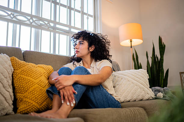 A woman with curly hair sits on a beige couch hugging her knees, looking out the window with a thoughtful expression, surrounded by cozy pillows and warm lighting.