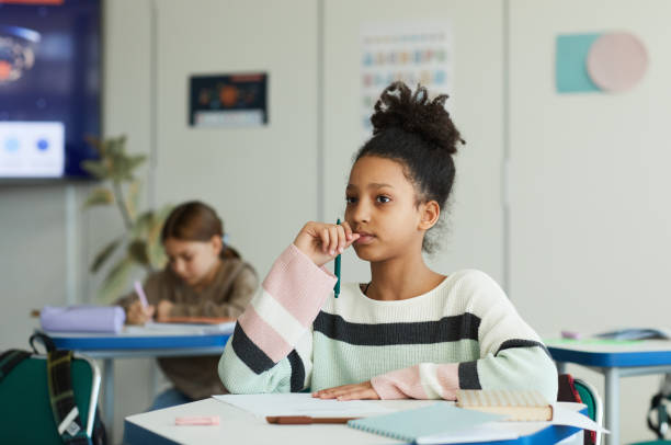 A preteen girl sitting in class with her chin resting on her hand, gazing off to the side with a bored and distant expression. She is surrounded by classmates but appears disengaged from the lesson.