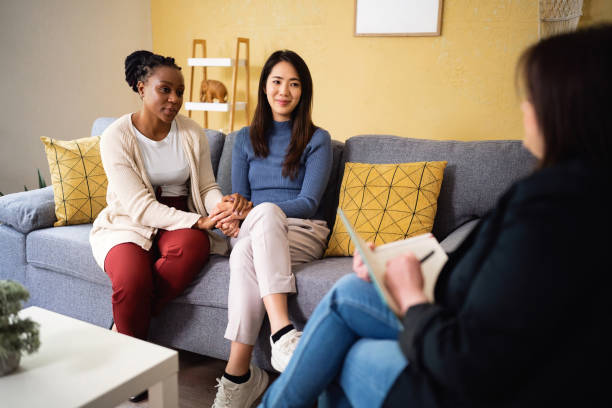 Two women sitting on a couch holding hands and speaking with a therapist, symbolizing emotional support and premarital counseling in a diverse relationship.
